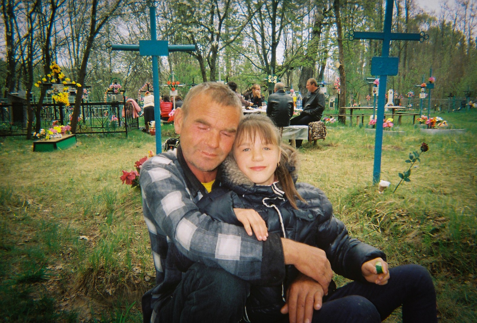 A father and daughter at a local cemetery during Easter celebrations. A table of villagers sit in the background eating and drinking